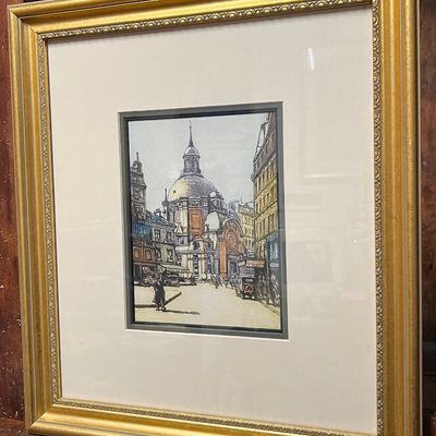 Framed architectural print depicting a European city street scene with a cathedral, housed in a double mat and ornate gold-finished frame.