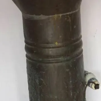 Antique copper or brass tall pitcher vessel with an arched top wire handle and side ceramic grip, displaying heavy age patina and verdigris.
