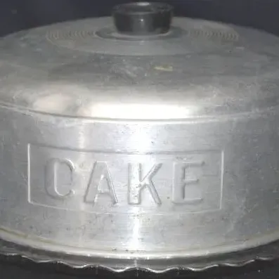 Vintage mid-century spun aluminum cake carrier with embossed 'CAKE' lettering and a black top knob, set on a scalloped glass base.