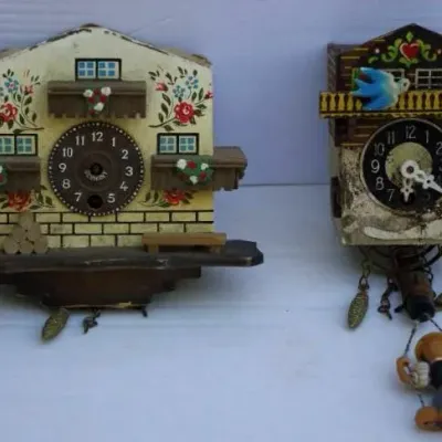 Two vintage German miniature cuckoo clocks featuring hand-painted chalet details, decorative pinecone weights, and swinging figures,...