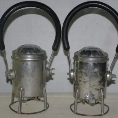 Pair of vintage railroad lanterns with metallic bodies, wire bases, and large black rubber-wrapped carrying handles.
