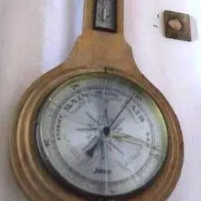 Vintage light wood banjo-style barometer featuring a central pressure gauge, upper thermometer, and lower secondary dial, showing visible...