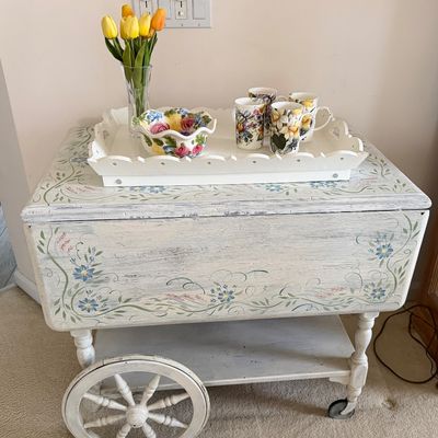 Sale Photo Thumbnail #2: Hand-painted white wooden tea cart featuring drop-leaf sides, floral motifs, and spoked wheels, display-styled with a tray, floral mugs, and a vase of tulips.