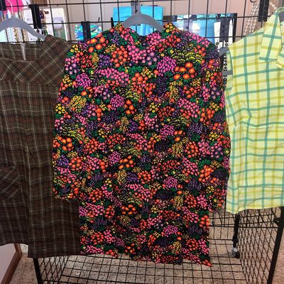 Trio of vintage 1960s to 1970s women's tops and dresses hanging on a display rack, including a brown plaid dress, a vibrant floral...