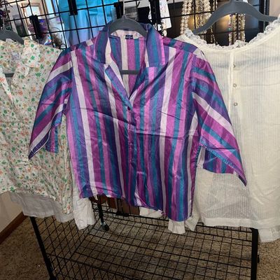 Trio of vintage women's blouses hanging on a rack, featuring a floral print sleeveless top, a bold purple and blue striped shirt,...