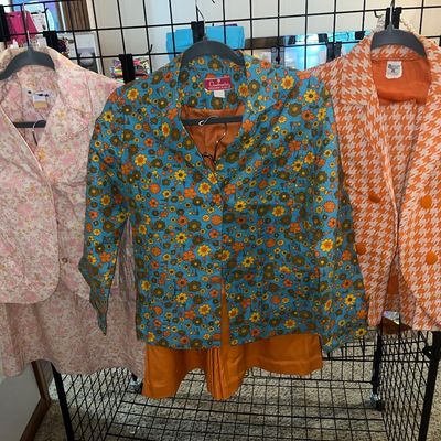 Trio of vintage 1970s women's skirt and shorts suits hanging on a wire display rack, including floral prints and an orange...