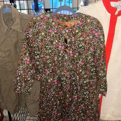 Trio of vintage garments on hangers, including a military shirt jacket, a floral pattern long-sleeve dress, and a white coat with red trim.