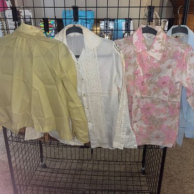 Selection of vintage women's blouses from the 1970s hanging on a display rack, including lace-trimmed, high-collar, floral, and...