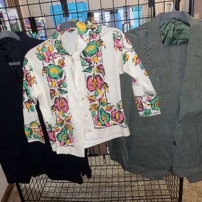 Trio of vintage apparel on hangers, including a black blazer, a colorful fruit-patterned button-up shirt, and a grey patterned men's...