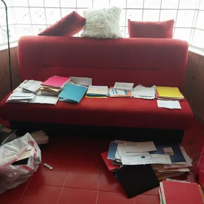 Sale Photo Thumbnail #28: Red armless sofa bed adorned with throw pillows, covered in stacks of sorted paper files and documents.