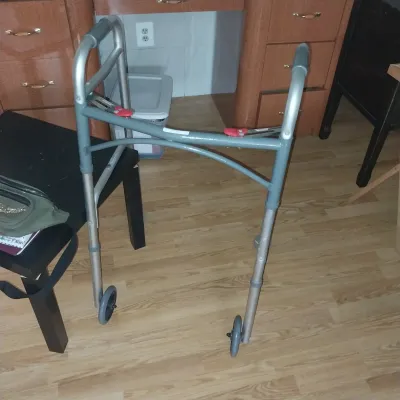 Sale Photo Thumbnail #24: Folding medical walker frame in gray aluminum, featuring front wheels, rubber hand grips, and red push-button release mechanisms.