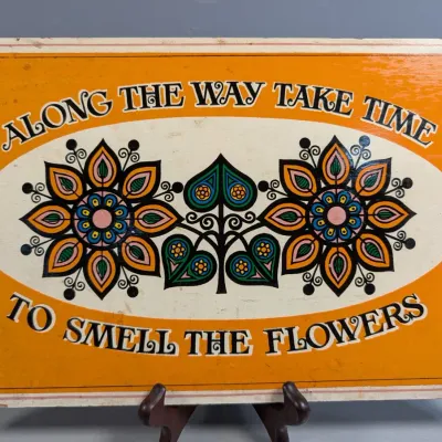Sale Photo Thumbnail #365: Vintage 1970s wooden wall plaque reading "Along the way take time to smell the flowers" with stylized floral illustrations on a bright orange background.