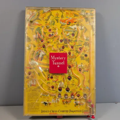 Sale Photo Thumbnail #168: Vintage Ideal's Cross Country Bagatelle Game featuring a yellow illustrated board, clear plastic cover, and central Mystery Tunnel box.