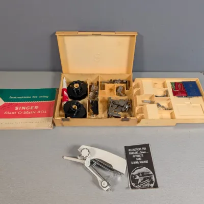 Sale Photo Thumbnail #368: Vintage Singer Slant-O-Matic 401 sewing machine accessory case with presser feet, bobbins, and original instruction booklet, alongside a portable handheld sewing device.