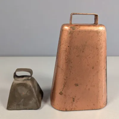 Sale Photo Thumbnail #173: Pair of vintage metal cowbells, featuring one large copper-toned bell and a smaller distressed iron bell, showing age-appropriate patina and wear.