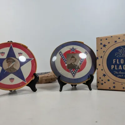 Sale Photo Thumbnail #77: Pair of World War II-era patriotic glass portrait plaques featuring military personnel, displayed on black stands next to an original Floral Plaques box.