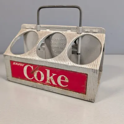 Sale Photo Thumbnail #129: Vintage Enjoy Coke aluminum six-pack bottle caddy featuring a wire carrying handle and embossed lettering, showing light wear.