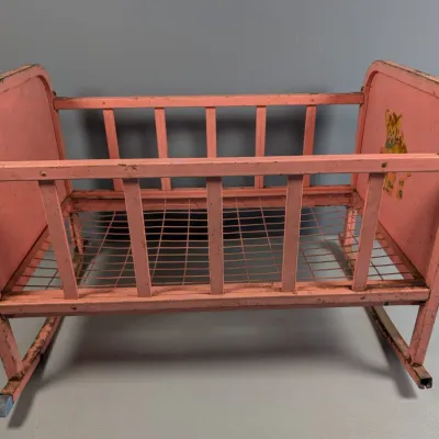 Sale Photo Thumbnail #172: Vintage pink metal doll crib or cradle with rocking base and wire spring bottom, featuring worn paint and a cartoon decal.