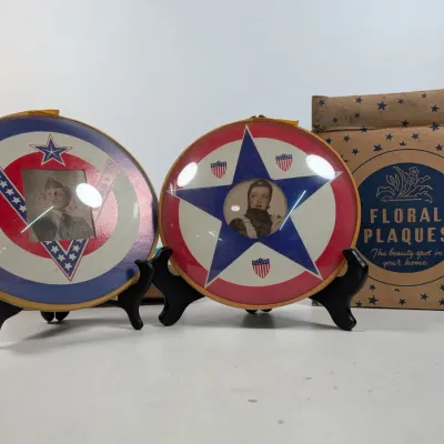 Sale Photo Thumbnail #70: Pair of World War II-era patriotic round convex glass photo plaques with red, white, and blue military motifs, displayed alongside an original 'Floral Plaques' cardboard box.