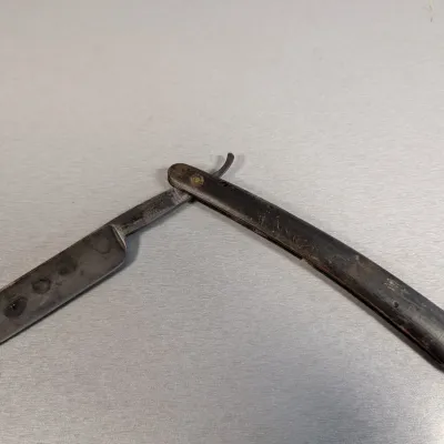 Sale Photo Thumbnail #392: Vintage straight razor featuring a steel blade with patina and a dark handle, showing signs of age and wear.