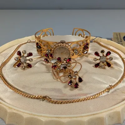 Sale Photo Thumbnail #378: Vintage Cort gold-tone women's cuff watch and matching rhinestone jewelry set, featuring red and clear stones including a necklace, brooch, and clip-on earrings displayed on a satin-lined tray.
