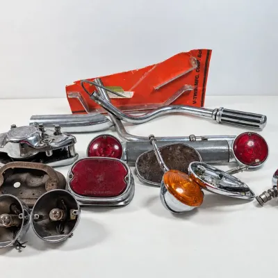 Sale Photo Thumbnail #64: Assortment of vintage motorcycle parts, including turn signal lights, tail light lenses, chrome handlebars, and mirrors, showing signs of wear and age.