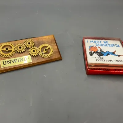 Sale Photo Thumbnail #298: Pair of vintage mid-century novelty items, including a wooden desk plaque with brass gears reading 'UNWIND!' and a boxed novelty tile reading 'I MUST BE SUCCESSFUL... I OWE EVERYBODY MONEY!'.