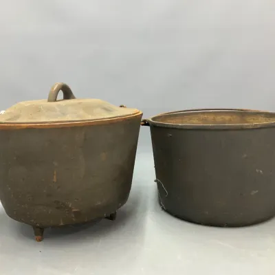 Sale Photo Thumbnail #264: Pair of antique cast iron cooking pots, including a three-legged Dutch oven with lid and a wire-handle kettle, showing heavy surface patina and age-related wear.