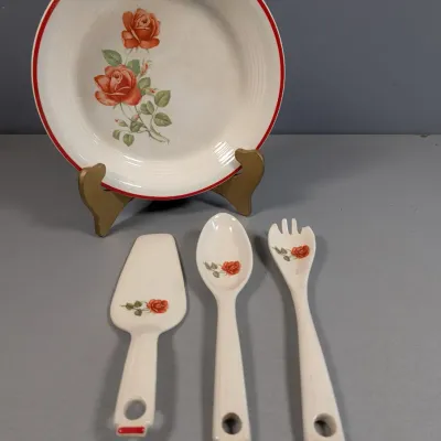 Sale Photo Thumbnail #179: Vintage ceramic serving set featuring a round serving bowl with a red rose motif and red rim trim, displayed on a wooden stand, accompanied by three matching ceramic serving utensils.