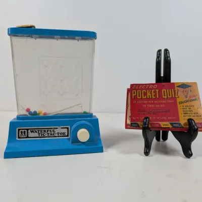 Sale Photo Thumbnail #422: Vintage Tomy Waterful Tic-Tac-Toe handheld game alongside an Electro Pocket Quiz matching game displayed on a stand.