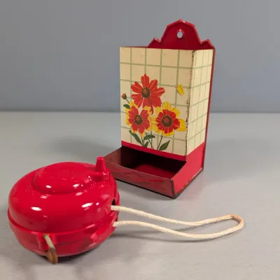 Sale Photo Thumbnail #330: Vintage red plastic string/twine dispenser alongside a vintage lithographed tin match holder featuring a floral design on a grid background.