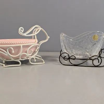 Sale Photo Thumbnail #136: Two sleigh-shaped decorative candy dishes, including a pink ceramic bowl in a white metal runner frame and a cut lead crystal sleigh on a dark wrought iron frame.