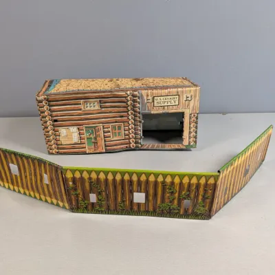 Sale Photo Thumbnail #447: Vintage Marx lithographed tin U.S. Cavalry Supply building with fold-out stockade fence, featuring detailed Western-style log cabin graphics.