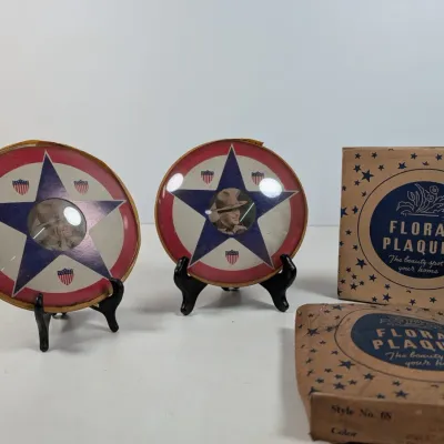 Sale Photo Thumbnail #69: Pair of vintage patriotic round wall plaques featuring stars, shields, and central portraits, displayed on black stands with original Floral Plaques boxes.