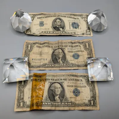 Sale Photo Thumbnail #18: Three vintage US silver certificate bills, including a 1953 $5 bill and two 1957 $1 bills, displayed alongside four clear cut-crystal paperweights.