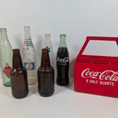 Sale Photo Thumbnail #47: Collection of vintage soda bottles including Coca-Cola, Pepsi, and Royal Crown Cola alongside a red plastic Coca-Cola six-pack carrier.