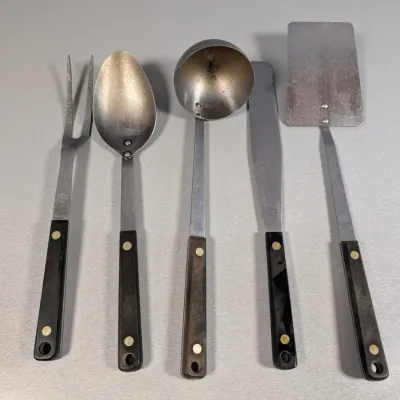 Sale Photo Thumbnail #437: Set of five vintage Flint stainless steel kitchen utensils featuring black composite handles with brass rivets. Includes a carving fork, serving spoon, ladle, frosting spatula, and turner.