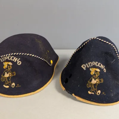 Sale Photo Thumbnail #349: Pair of vintage Disney Pinocchio navy felt beanie hats with yellow trim and twisted cord detail, showing heavy wear and moth damage.