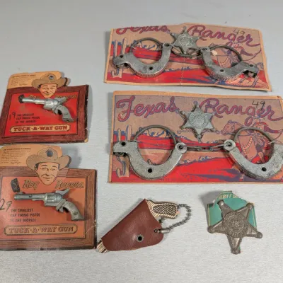Sale Photo Thumbnail #317: Collection of vintage western toy accessories, including Roy Rogers miniature cap pistols on original cardstock packaging, Texas Ranger play handcuffs, a small holster, and a sheriff badge.
