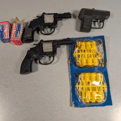 Sale Photo Thumbnail #309: Collection of vintage toy cap guns alongside boxes of Kilgore perforated roll caps and a carded package of yellow plastic play bullets.