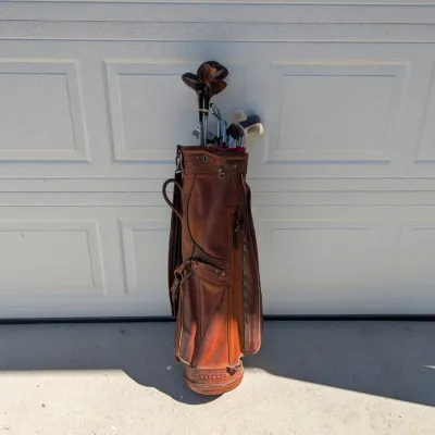 Sale Photo Thumbnail #249: Vintage brown leather golf cart bag filled with an assortment of classic wooden woods and iron golf clubs.