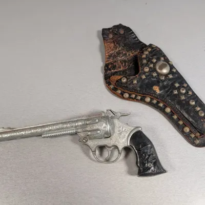 Sale Photo Thumbnail #313: Vintage Hubley Kit Carson toy cap gun revolver with scrollwork engraving and black plastic grips, accompanied by a studded black leather holster.