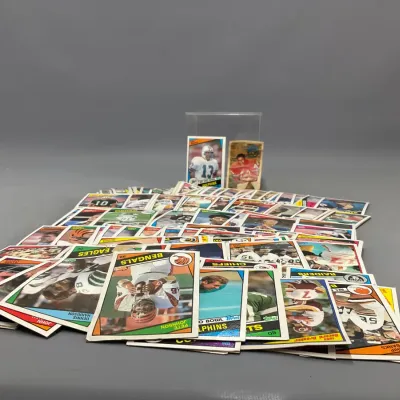 Sale Photo Thumbnail #405: Large collection of vintage football trading cards from the 1970s and 1980s, primarily Topps, scattered across a surface with two highlighted cards in the background.