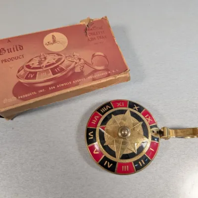 Sale Photo Thumbnail #440: Vintage Guild Products SunDial roulette ash tray in brass and enamel, accompanied by its original box showing wear.