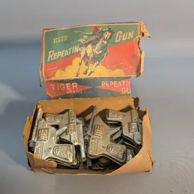Sale Photo Thumbnail #318: Vintage box of Tiger Brand tin metal repeating toy cap guns, made in Japan, featuring colorful western graphics on the damaged original packaging.