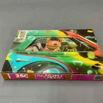 Sale Photo Thumbnail #230: Vintage Milton Bradley 250-piece jigsaw puzzle featuring The Muppet Movie, depicting Kermit the Frog and Fozzie Bear driving a car, showing shelf wear and tape residue on the box.