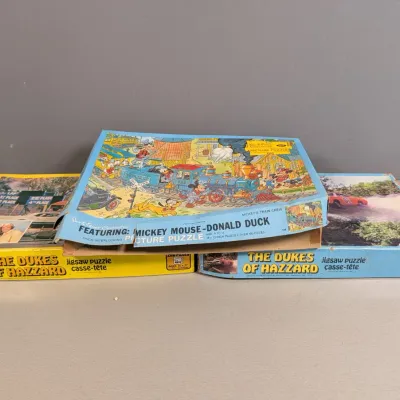 Sale Photo Thumbnail #194: Collection of three vintage 1970s–1980s pop culture jigsaw puzzles, featuring a Walt Disney Mickey Mouse and Donald Duck train scene alongside two The Dukes of Hazzard puzzle boxes showing wear.