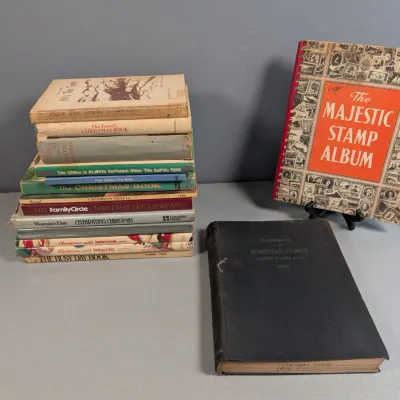 Sale Photo Thumbnail #449: Collection of vintage books including a stack of Christmas-themed hardcovers, a 1953 US Navy Hospital Corps handbook, and 'The Majestic Stamp Album'.
