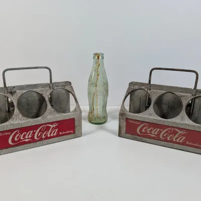 Sale Photo Thumbnail #46: Pair of vintage aluminum Coca-Cola six-bottle metal carriers with wire handles alongside a green glass Coca-Cola contour bottle.