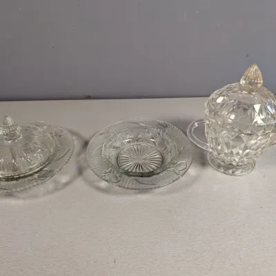 Sale Photo Thumbnail #177: Trio of vintage pressed clear glass items, including a covered butter dish, an additional saucer or bowl, and a diamond-point pattern covered sugar bowl.